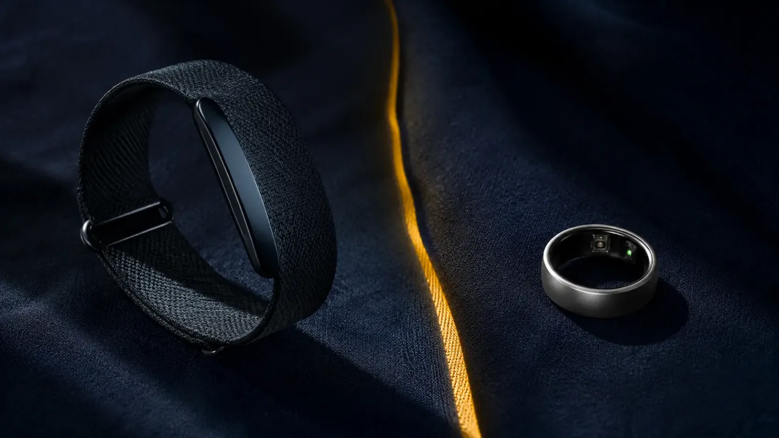 A black screenless fitness band and a titanium smart ring facing each other on navy fabric, separated by a thin mustard line of light